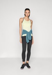Lichtgeel sporttanktop met zwarte patroonstraps, zwarte aansluitende legging en een teal shirt geknoopt om de taille. Zwarte sportschoenen.