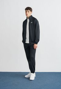 Μαύρο φόρμα με ζακέτα με φερμουάρ και ταιριαστό παντελόνι, συνδυασμένα με λευκό μπλουζάκι και λευκά αθλητικά παπούτσια. Λείο υλικό, άνετη εφαρμογή.