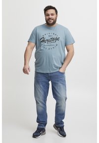 Gaiši zils kokvilnas T-krekls ar melnu grafisku tekstu, kas saka "Original No. 1 Heritage Blend". Valkāts kopā ar ziliem džinsiem un melnām sporta apaviem.