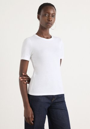 Jeune femme aux cheveux courts portant des boucles d'oreilles créoles, un t-shirt blanc ajusté à manches courtes et un jean bleu foncé, debout avec un bras croisé.