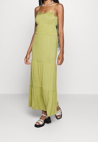 Robe maxi vert olive avec des bretelles froncées, taille froncée, jupe à volants et texture douce. Associée à des sandales à talons carrés noires.