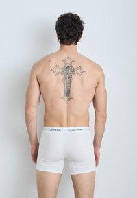 Boxeri din bumbac alb cu un chenar negru "Calvin Klein", purtați de un bărbat care își arată un tatuaj detaliat pe spate, pe un fundal deschis.
