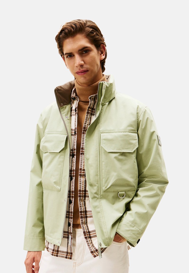 Chaqueta verde claro, resistente al agua, con un cuello alto, dos bolsillos frontales y un forro interior marrón en contraste. Lleva puesta sobre una camisa con estampado.