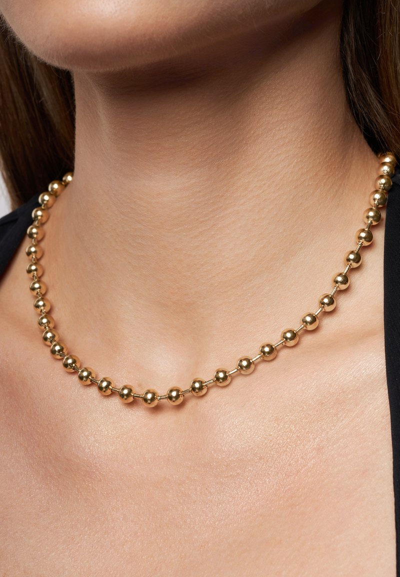 Gold bead necklace with alternating spherical beads and small connectors, resting on the neck, with a smooth and shiny texture.