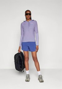 Šviesiai violetinis ilgomis rankovėmis megztinis su gaubtu ir pusiau užtrauktuku, mėlynos spalvos šortai, pilki sportbačiai ir juoda kuprinė. Lygi tekstūra ir laisvas siluetas.