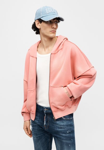 Ροζ απαλή ζακέτα με φερμουάρ και άνετη εφαρμογή, με κουκούλα, μπροστινές τσέπες και φθαρμένες λεπτομέρειες. Συνδυασμένη με μπλε τζιν παντελόνι.