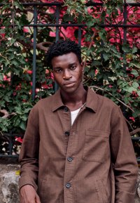 Young man with short curly hair wearing a brown buttoned jacket stands against a black fence with green and red foliage.