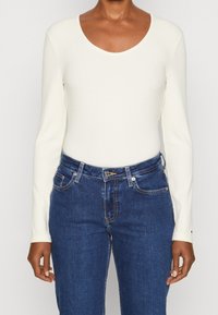 Long-sleeve, ribbed bodysuit in off-white, with a scoop neckline. Paired with blue denim jeans featuring traditional pockets and stitching.