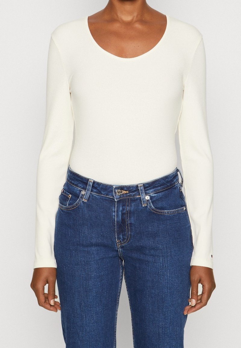 Long-sleeve, ribbed bodysuit in off-white, with a scoop neckline. Paired with blue denim jeans featuring traditional pockets and stitching.