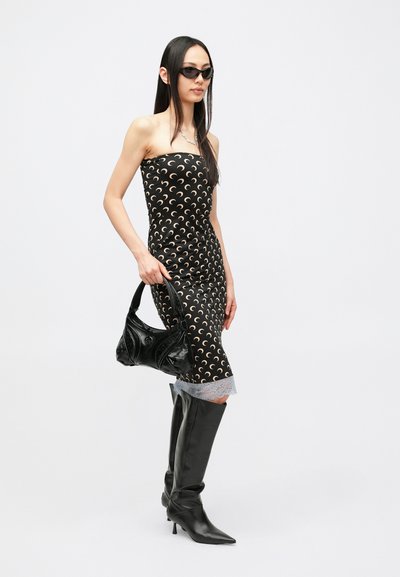 Czarna sukienka bez ramiączek z kremowym wzorem w kształcie półksiężyców, z warstwą koronki u dołu. Modelka trzyma czarną torebkę o fakturze; ma na sobie czarne kozaki sięgające kolan.