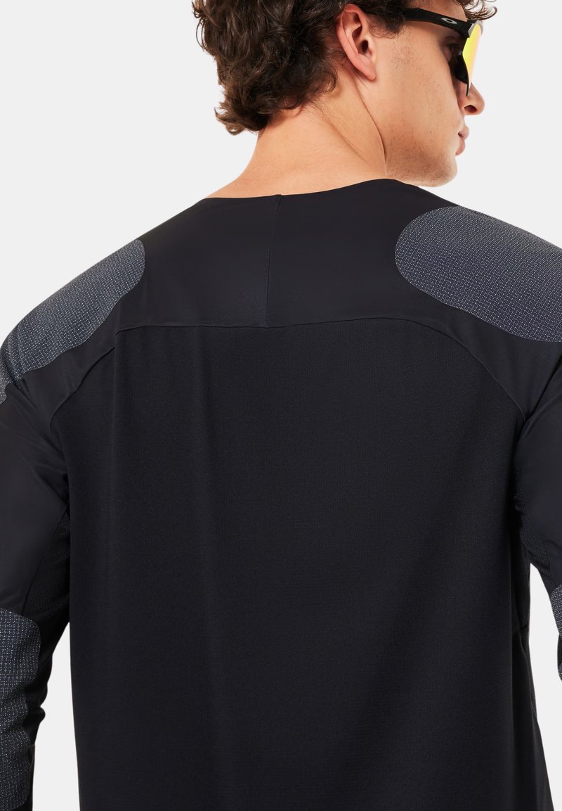 Homme portant un haut de sport noir et gris à manches longues, vu de dos, avec des cheveux courts et bouclés et des lunettes de soleil noires aux verres jaunes.