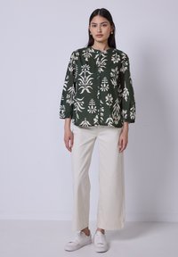 Blusa con stampa floreale verde, con bottone frontale, maniche a tre quarti e polsini arricciati, abbinata a pantaloni ampi di colore chiaro e sneakers bianche.