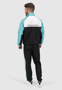 Zwarte en teal trainingspak met een witte bovenkant en het Sergio Tacchini-logo. Bevat elastische manchetten en broek met trekkoord, met retro ontwerpaccenten.