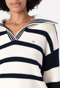 Pulover tricotat cu dungi verticale alb-negru și crem, cu guler în formă de V cu textură ribbed și un mic logo pe piept.