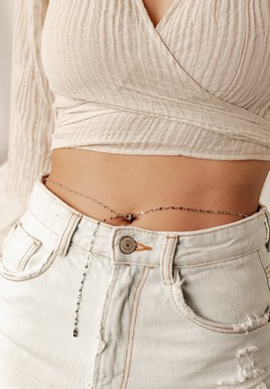 Ceinture en chaîne argentée avec des maillons complexes et un petit fermoir rond, portée sur un short en denim blanc taille haute avec des bords effilochés et des coutures orange.
