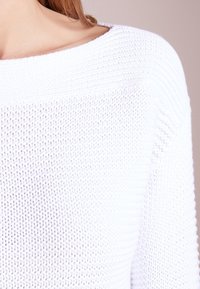Femme portant un pull en tricot texturé blanc avec un décolleté large, révélant une partie du cou et de l'épaule.