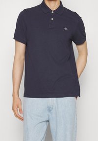 Homme portant un polo bleu marine à manches courtes avec des boutons et un jean bleu clair, se tenant devant un fond uni.