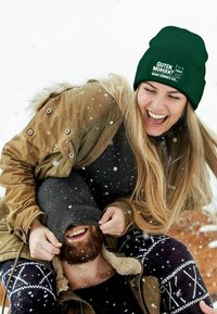 Green knitted beanie with text and graphic, worn by a woman with long hair, in a snowy outdoor setting, with a man partially obscured below.
