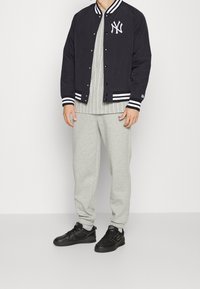 Marinblå basebolljacka med vita ränder och logotyp, grårandig skjorta, grå joggingbyxor och svarta sneakers på en enfärgad bakgrund.