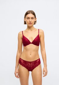 Νεαρή γυναίκα που φοράει κλασικό κόκκινο δαντελένιο σουτιέν και σλιπ, στέκεται μπροστά σε απλό φωτεινό φόντο, κοιτάζοντας ευθεία με ουδέτερη έκφραση.