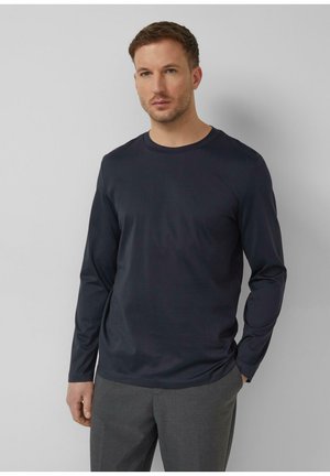 Uomo con capelli castani corti, che indossa una camicia blu scuro a maniche lunghe e pantaloni grigi, in piedi davanti a uno sfondo semplice di colore grigio chiaro.