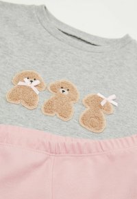 Graues Shirt mit drei beigen Pudel-Applikationen, die pinke Schleifen tragen. Der Boden hat einen weichen, pinken Gummibund.