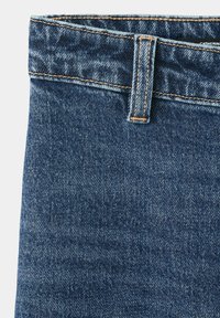 Gros plan sur la ceinture d’un jean en denim bleu avec une boucle de ceinture et des coutures orange visibles sur un fond clair.