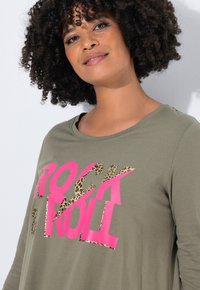 Camiseta verde oliva con cuello redondo, que presenta un texto en negrita en rosa "ROCK" y "ROLL" en estampado de leopardo. Tejido suave y ligero.