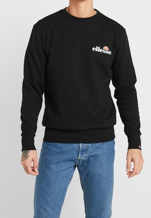 Felpa nera con scollo rotondo e polsini a costine, con logo Ellesse bianco sul petto. Indossata con jeans blu.