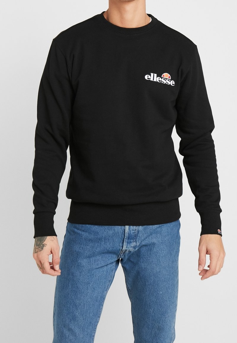 Felpa nera con scollo rotondo e polsini a costine, con logo Ellesse bianco sul petto. Indossata con jeans blu.