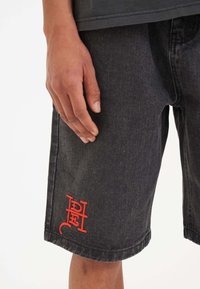 Black denim shorts with an embroidered red logo detail, featuring a straight cut and visible stitching, shown worn with a hand resting on them.