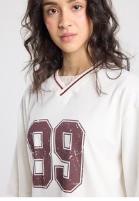 Wit oversized T-shirt met een vintage "89" grafiek in kastanjebruin, v-hals ontwerp en kastanjebruine bies. Zachte stoftextuur, casual pasvorm.