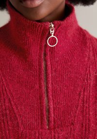 Roter Wollpullover mit hohem Kragen und einem halben Reißverschluss mit rundem Ziehgriff. Die Textur ist gerippt mit ausgeprägten vertikalen Mustern.