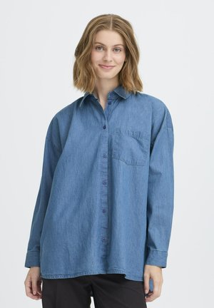 Junge Frau mit schulterlangem Haar, die ein übergroßes, blaues Jeanshemd und schwarze Hosen trägt und sanft lächelt.