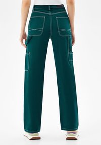 Nainen, jolla on yllään tummanvihreät leveälahkeiset cargo-housut, joissa on suuret takataskut ja sivutaskut, yhdistettyinä valkoisiin tennareihin, katsottuna takaa.