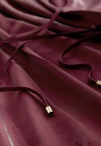 Bordó szövet sima textúrával, amelyen húzózsinórok és arany fémes végződések találhatók. A szövet lágyan omlik, finom redőket alkotva.