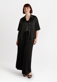 Camisa corta de satén negro, superpuesta a una blusa a juego, combinada con pantalones de pierna ancha y sandalias negras con adornos.