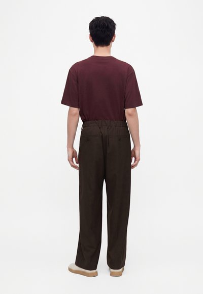 Homme debout, de dos, portant un t-shirt rouge foncé, un pantalon ample marron foncé avec des poches arrière, et des chaussures blanches sur un fond uni.