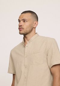 Chemise beige à manches courtes en tissu texturé, avec un design à boutons, un col mandarin et une poche sur la poitrine.