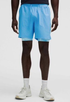 Männlich, trägt hellblaue, geschichtete Sportsshorts, weiße Kniestrümpfe und weiße Laufschuhe und steht vor einem schlichten Hintergrund.