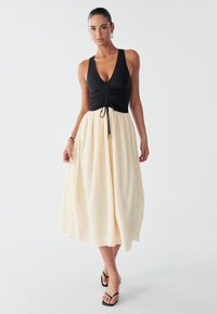 Svart ärmlös topp med rynkad design, som övergår i en krämfärgad texturerad maxikjol. Bärs med svarta sandaler.