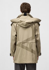 Beige huva jacka med cinchad midja, tillverkad av lätt material. Har en stor, subtil logotypdesign på baksidan och svarta accenter.