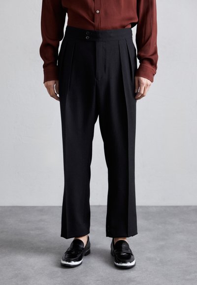 Pantalons plissés noirs à taille haute, dotés de deux boutons, associés à des mocassins noirs et à une chemise bordeaux. Texture de tissu lisse.