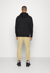 Sudadera negra con bolsillo canguro, pantalones beige ajustados y zapatillas negras. La tela parece suave; el conjunto presenta un ajuste relajado.