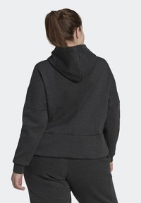 Femme avec une queue de cheval brune portant un sweat à capuche gris foncé et un pantalon de survêtement assorti, vue de dos sur un fond uni.