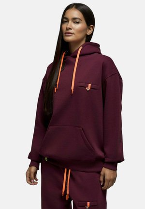 Femme portant un sweat à capuche bordeaux et un pantalon assorti avec des cordons orange et de petites poches boutonnées, debout devant un fond uni.