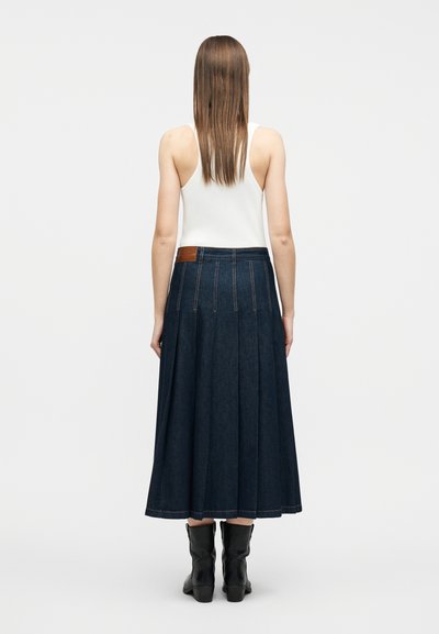 Γυναίκα με ίσια καστανά μαλλιά, που φοράει λευκή αμάνικη μπλούζα, σκούρα μπλε ριχτή τζιν φούστα και μαύρες μπότες μέχρι τη μέση της γάμπας, εμφανιζόμενη από πίσω.