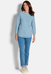 Hellblaues langärmliges Sweatshirt mit weitem Ausschnitt, kombiniert mit hellen Jeans und beigen Turnschuhen. Glatte Textur, lässige Passform.