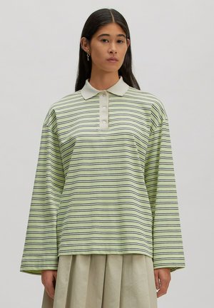 Woman with long dark hair wearing a light green and white striped long-sleeve polo shirt and a beige pleated skirt against a plain background.