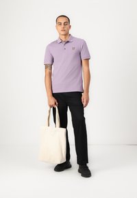 Lilla polo skjorte med tre knapper og gult logo, sammen med svarte bukser og en beige kanvas tote bag. Svarte joggesko brukt.